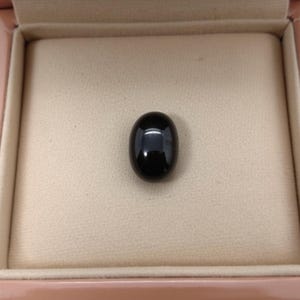 Könnte beinhalten: Ein polierter, oval geformter schwarzer Edelstein, präsentiert in einer hellbraunen Schmuckschatulle. Der Edelstein ist auf einer beigefarbenen Samtoberfläche in der Schatulle zentriert. Die Schatulle hat eine glänzende, hellbraune Oberfläche.