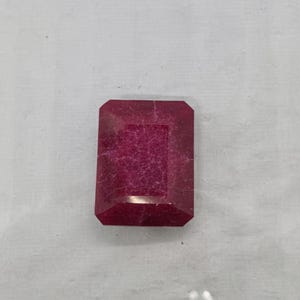 Könnte beinhalten: Ein facettierter, rechteckiger Rubin-Edelstein mit tiefroter Farbe. Der Edelstein hat eine leicht strukturierte Oberfläche und ein poliertes, reflektierendes Aussehen. Der Schliff ist ein Smaragdschliff, mit rechteckiger Form und abgeschrägten Ecken.