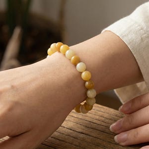 Könnte beinhalten: Ein Armband mit runden, gelben und cremefarbenen Perlen. Das Armband wird am Handgelenk getragen und zeigt die verschiedenen Farbtöne der Perlen. Der Hintergrund umfasst eine Holzoberfläche und einen Blick auf eine Pflanze.
