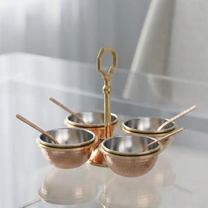 Op de afbeelding: Een kruidenserveerset bestaande uit vier kleine, ronde, gehamerde koperen kommen met zilverkleurige binnenkanten. Elke kom bevat een bijpassende metalen lepel. De kommen zijn gerangschikt rond een centrale verticale messing standaard.