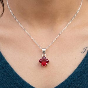 Könnte beinhalten: Eine silberne Halskette mit einem quadratischen, tiefroten Edelsteinanhänger. Der Anhänger ist in eine silberne Fassung gefasst und hängt an einer filigranen Silberkette. Die Halskette wird von einer Person mit einem teal-farbenen Oberteil getragen.