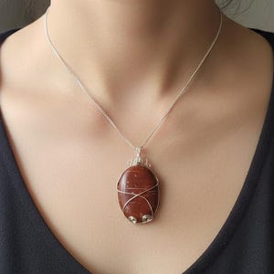 Könnte beinhalten: Eine silberne Halskette mit einem Anhänger aus rotbraunem Stein. Der ovale Stein ist mit Silberdraht umwickelt und hängt an einer zarten Silberkette. Der Anhänger ist ein einzigartiges Schmuckstück mit polierter Oberfläche.