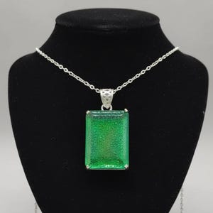 Könnte beinhalten: Eine silberne Halskette mit einem rechteckigen, smaragdgrünen Anhänger. Der Anhänger hat ein glitzerndes, strukturiertes Aussehen und hängt an einer zarten Kette. Die Halskette wird auf einer schwarzen Schaufensterpuppe präsentiert.