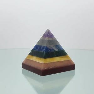 May include: A multi-colored pyramid-shaped decorative object. The pyramid features layers of various colors, including brown, yellow, blue, and purple. The pyramid is sitting on a glass surface.