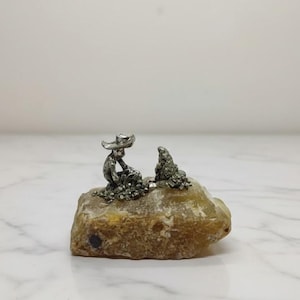 Könnte beinhalten: Kleine silberne Figur einer Person mit Hut, die auf einem hellbraunen, strukturierten Felsen sitzt, mit einer weiteren Figur daneben. Die Figur ist wahrscheinlich ein dekorativer Gegenstand.