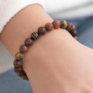 Könnte beinhalten: Ein Perlenarmband mit Natursteinperlen in Braun-, Rot- und Gelbtönen. Das Armband wird am Handgelenk getragen und hebt die erdigen Farbtöne und Texturen der Edelsteine hervor. Die Perlen haben einen Durchmesser von etwa 0,8 cm.