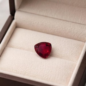 Könnte beinhalten: Ein dreieckiger, tiefroter Edelstein liegt in einer offenen, braunen Schmuckschatulle mit cremefarbenem Innenraum. Der Edelstein ist der Blickfang und zeigt seine leuchtende Farbe und Form. Die Schatulle befindet sich vor einem weichen, neutralen Hintergrund.