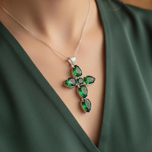 Puede incluir: Un collar de plata con un colgante de cruz. La cruz está adornada con grandes piedras preciosas verdes en forma de pera. El collar se lleva sobre una blusa verde oscuro.