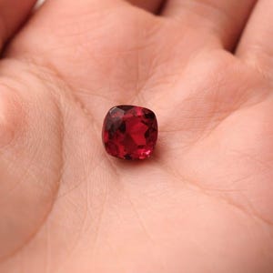 Könnte beinhalten: Ein facettierter, kissenförmiger, tiefroter Edelstein. Der Edelstein wird in einer offenen Handfläche gehalten, wodurch seine leuchtende Farbe und reflektierenden Oberflächen zur Geltung kommen. Die Form des Steins ist quadratisch mit abgerundeten Ecken.