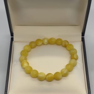 Könnte beinhalten: Ein gelbes Perlenarmband, präsentiert in einer offenen, dunkelbraunen Schmuckschatulle mit cremefarbenem Innenraum. Das Armband besteht aus runden, hellgelben Perlen. Die Schatulle ist geöffnet und zeigt das Armband auf einer weichen, gepolsterten Oberfläche.