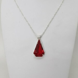 Könnte beinhalten: Eine silberne Halskette mit einem großen, facettierten, roten Anhänger. Der Anhänger hat eine tiefrote Farbe und eine geometrische, dreieckige Form. Die Halskette wird auf einer weißen Schaufensterpuppe präsentiert.