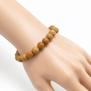Könnte beinhalten: Ein hellbraunes Holzperlenarmband. Das Armband besteht aus runden, polierten Perlen, die aneinandergereiht sind. Die Perlen haben ein natürliches Holzmaserungsmuster und werden am Handgelenk getragen.