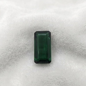 Könnte beinhalten: Ein rechteckiger, smaragdgrüner Edelstein mit Facettenschliff. Der Edelstein ist vor einem weichen, weißen Hintergrund platziert. Die tiefgrüne Farbe und der präzise Schliff des Edelsteins werden hervorgehoben.
