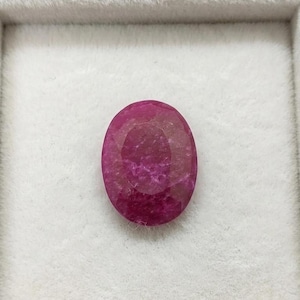Könnte beinhalten: Ein facettierter, oval geformter Rubin-Edelstein. Der Edelstein hat eine tiefe, gesättigte, rötlich-rosa Farbe, mit sichtbaren inneren Einschlüssen. Der Rubin liegt vor einem weißen Hintergrund.
