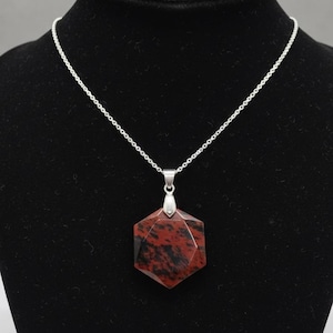 Könnte beinhalten: Halskette mit silberner Kette und geometrischem Anhänger. Der Anhänger hat eine sechseckige Form mit einem roten und schwarzen Muster. Die Halskette wird auf einer schwarzen Schaufensterpuppe präsentiert.