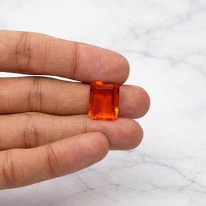 Könnte beinhalten: Ein leuchtend orangefarbener, rechteckiger Edelstein mit Facettenschliff. Der Edelstein wird in einer Hand gehalten, wodurch seine Farbe und Klarheit hervorgehoben werden. Der Stein hat eine rechteckige Form und scheint ein Edelstein oder ein dekoratives Element zu sein.