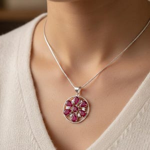 Könnte beinhalten: Eine silberne Halskette mit Anhänger, die ein kreisförmiges Design aufweist. Der Anhänger ist mit tropfenförmigen, tiefroten Edelsteinen und kleineren weißen Edelsteinen besetzt. Die Halskette hängt an einer filigranen Silberkette.