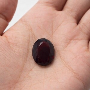 Könnte beinhalten: Ein facettierter, oval geformter, tiefroter Edelstein. Der Edelstein wird in einer offenen Handfläche gehalten, wodurch seine polierte Oberfläche und seine satte Farbe zur Geltung kommen. Der Schliff des Steins lässt das Licht reflektieren und erzeugt einen funkelnden Effekt.