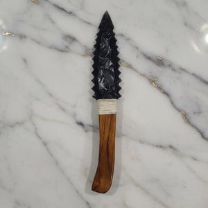 Könnte beinhalten: Ein handgefertigter Obsidian-Pfeilspitze mit gezackter Kante, montiert auf einem Holzgriff. Der Griff ist mit einer Naturfaserschnur umwickelt. Die Pfeilspitze ist tiefschwarz, das Holz hat einen warmen, braunen Farbton. Ein dekoratives Objekt.