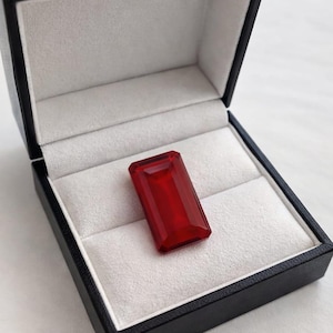 Könnte beinhalten: Ein rechteckiger, facettierter, tiefroter Edelstein liegt in einer schwarzen Schmuckschatulle mit weißem Innenfutter. Der Edelstein ist der Blickfang und zeigt seine satte Farbe und geometrische Form. Die Schatulle ist geöffnet.