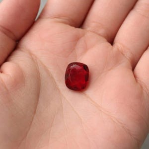 Könnte beinhalten: Ein facettierter, kissenförmiger, tiefroter Edelstein liegt in der Handfläche. Der Edelstein hat eine polierte Oberfläche und reflektiert das Licht, wodurch seine satte Farbe und Klarheit zur Geltung kommen. Der Hintergrund ist ein neutrales Grau.