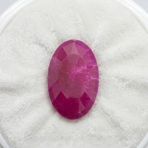 Könnte beinhalten: Ein facettierter, oval geformter Rubin-Edelstein. Der Edelstein hat eine tiefe, lebendige rosa-rote Farbe und ist vor einem weißen, strukturierten Hintergrund platziert. Der Schliff des Steins erzeugt mehrere reflektierende Oberflächen.