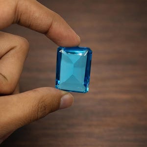 Könnte beinhalten: Ein rechteckiger, facettierter, hellblauer Edelstein, der zwischen Fingern gehalten wird. Der Edelstein hat einen Brillantschliff mit einer zentralen Facette und ist vor einem dunklen Holzmaserungshintergrund platziert. Die Farbe des Edelsteins ist ein lebendiges, klares Blau.
