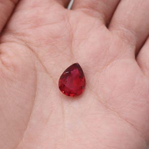 Könnte beinhalten: Ein facettierter, tropfenförmiger, tiefroter Edelstein. Der Edelstein wird in einer Hand gehalten, um seine brillante Farbe und seinen Schliff zu zeigen. Die Facetten des Steins fangen das Licht ein und erzeugen einen funkelnden Effekt.