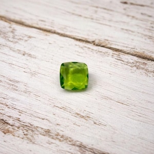 Könnte beinhalten: Ein quadratisch geschliffener, durchscheinender, limettengrüner Edelstein. Der Edelstein hat eine facettierte Oberfläche, die das Licht einfängt und reflektiert. Der Stein liegt auf einer verwitterten, hellen Holzoberfläche, was einen Kontrast in Texturen und Farben erzeugt.