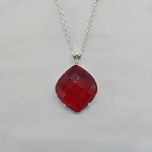 May include: A silver necklace with a large, faceted, red gemstone pendant. The pendant is a square shape with rounded corners. The chain is a delicate silver rope style.