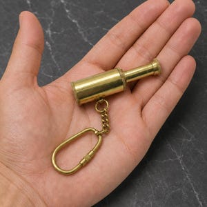 Könnte beinhalten: Ein Miniatur-Teleskop-Schlüsselanhänger aus Messing mit einem zylindrischen Körper, einem konischen Okular und einer strukturierten Oberfläche. Das Teleskop ist an einer kleinen Metallkette befestigt, die mit einem ovalen Messingkarabiner verbunden ist.