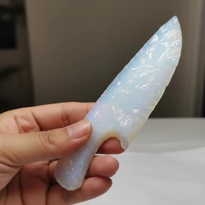 Könnte beinhalten: Ein durchscheinendes, opalisierendes Kristallmesser mit gebogenem Griff. Der Kristall hat ein glattes, poliertes Aussehen mit subtilen inneren Mustern. Das Messer wird in einer Hand gehalten, um seine Größe und Form zu zeigen. Die Gesamtästhetik ist natürlich und organisch.