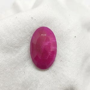 Könnte beinhalten: Ein leuchtender, oval geformter Edelstein mit einem tiefen Magenta-Pink-Farbton und einer facettierten Oberfläche. Der Stein hat ein poliertes Finish, das das Licht über seine zahlreichen geometrischen Schliffe reflektiert und eine durchscheinende Qualität zeigt.