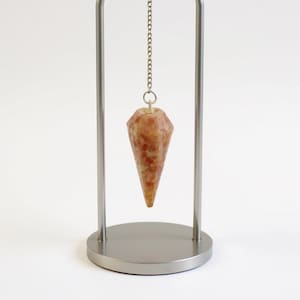 Könnte beinhalten: Ein Kristallpendel mit facettiertem, kegelförmigem Design in Orange- und Brauntönen, das an einer Kette an einem silbernen Metallständer hängt. Der Ständer hat eine kreisförmige Basis und zwei vertikale Stützen.