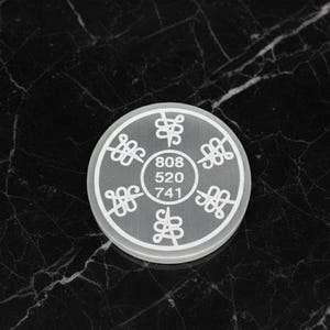 Könnte beinhalten: Ein runder, durchscheinend weißer Anhänger mit einer Reihe stilisierter Symbole und den Zahlen 808, 520 und 741 in der Mitte. Der Anhänger befindet sich vor einem dunklen, marmorierten Hintergrund.