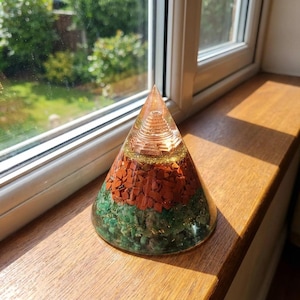 May include: A clear pyramid-shaped object with layers of red, green, and gold-colored materials. The pyramid sits on a wooden surface near a window. The top of the pyramid has a spiral design.