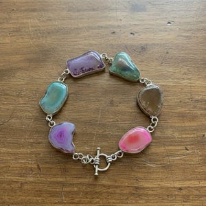 Könnte beinhalten: Ein silbernes Armband mit unregelmäßig geformten, farbenfrohen Achatsteinen. Die Steine sind in den Farbtönen Lila, Grün, Rosa und Braun gehalten. Das Armband hat einen Knebelverschluss.