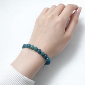 Könnte beinhalten: Ein blau-grünes Perlenarmband wird am Handgelenk getragen. Das Armband besteht aus runden, polierten Perlen. Das Handgelenk befindet sich vor einem weißen Hintergrund. Das Armband ist ein Schmuckstück.
