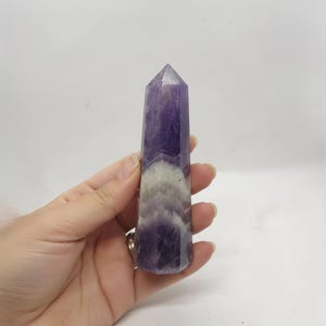 Könnte beinhalten: Ein polierter Amethyst-Kristall-Obelisk. Der lila Kristall hat eine spitze Spitze und ein geschichtetes Aussehen mit weißen und lila Bändern. Der Kristall wird in einer Hand vor weißem Hintergrund gehalten.