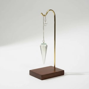 Könnte beinhalten: Ein Pendel mit einer klaren, facettierten Kristallspitze, die an einem goldfarbenen Metallständer aufgehängt ist. Der Ständer hat einen dunklen Holzsockel und eine gebogene Oberseite. Eine kleine Kette und eine Perle akzentuieren den Kristall.