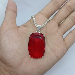 Könnte beinhalten: Eine silberne Halskette mit einem großen, rechteckigen, facettierten roten Anhänger. Der Anhänger ist in einen silbernen Rahmen gefasst und hängt an einer silbernen Kette. Der Edelstein hat eine tiefe, satte Farbe und eine polierte Oberfläche.
