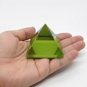Könnte beinhalten: Eine transparente, limettengrüne Pyramide mit spitzer Spitze. Die Pyramide wird in einer offenen Hand vor weißem Hintergrund gehalten. Die geometrische Form der Pyramide ist klar definiert, mit sichtbaren Facetten.