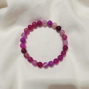 Könnte beinhalten: Ein Armband aus rosa- und lilafarbenen Edelsteinperlen. Die runden Perlen sind in verschiedenen Schattierungen von Magenta, Fuchsia und Lavendel gehalten. Das Armband liegt auf einem weißen Hintergrund.