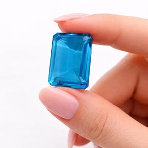 Könnte beinhalten: Ein großer, rechteckiger, facettierter, leuchtend blauer Edelstein. Der Edelstein wird zwischen zwei Fingern vor einem weißen Hintergrund gehalten. Die Facetten des Edelsteins reflektieren das Licht und erzeugen einen funkelnden Effekt. Der Edelstein ist ein lebendiges, auffälliges Stück.