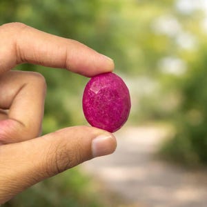 Könnte beinhalten: Ein leuchtender, oval geformter, facettierter Edelstein in einem tiefen Magentarosa. Der Stein weist eine texturierte, mehrfach facettierte Oberfläche auf, die das Licht einfängt und eine satte, gesättigte Farbe in seiner abgerundeten Form zeigt.