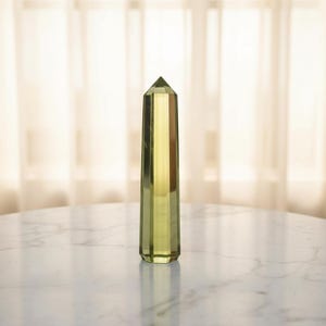 Könnte beinhalten: Ein hoher, facettierter, gelb-grüner Kristallobelisk. Der Kristall hat eine spitze Spitze und eine rechteckige Basis. Der Obelisk wird auf einem weißen Marmortisch mit einem weichen, neutralen Hintergrund präsentiert.