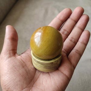 Può includere: Una sfera lucida giallo dorato poggia su una base cilindrica di colore chiaro. La sfera ha una finitura lucida, che riflette la luce. La base ha un aspetto strutturato, simile al legno. L'oggetto è tenuto in una mano aperta.