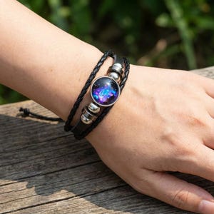 Könnte beinhalten: Schwarzes Lederarmband mit geflochtenem Band und rundem, galaktischem Mittelstück. Das Mittelstück zeigt eine lebendige Darstellung von Blau-, Lila- und Weißtönen, eingefasst in eine silberfarbene Lünette. Das Armband wird mit einer schwarzen Kordel befestigt.