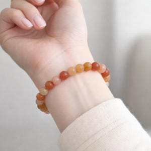 Könnte beinhalten: Ein Armband mit orangefarbenen, pfirsichfarbenen und rot getönten Perlen. Das Armband wird am Handgelenk getragen und zeigt die verschiedenen Farben der Perlen. Der Schmuck ist ein schlichtes, elegantes Stück. Die Perlen haben einen Durchmesser von etwa 0,8 cm.