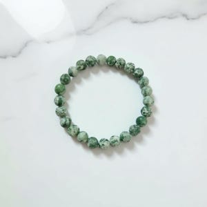 Könnte beinhalten: Armband aus grünen und weißen, gesprenkelten Perlen. Die runden Perlen sind zu einem Armband aufgereiht. Die Perlen haben einen Durchmesser von etwa 0,8 cm. Das Armband wird auf einer weißen Marmoroberfläche präsentiert.